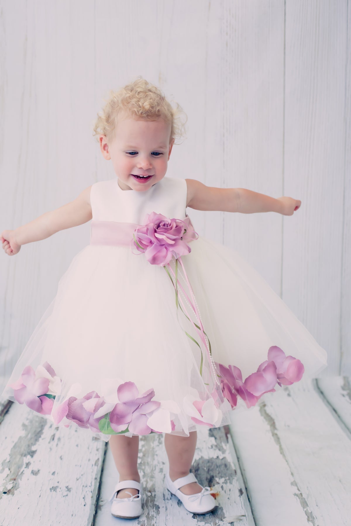 White satin baby dress with flower petal design and organza sash, by Kid's Dream, shown with delicate fabric details and floral accents.