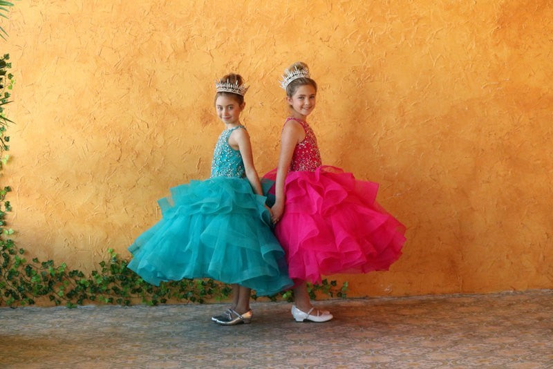Two girls in colorful dresses and tiaras standing against an orange wall.